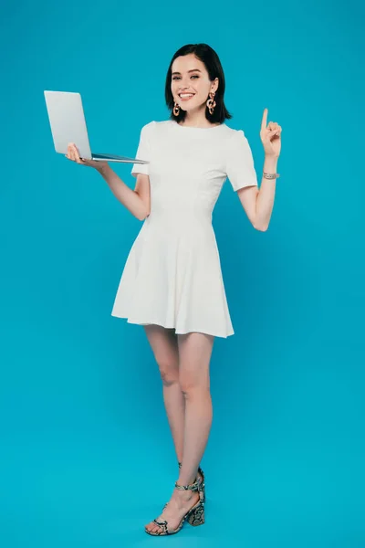 Повний вигляд усміхненої елегантної жінки, що тримає ноутбук і показує жест ідеї ізольовано на синьому — стокове фото