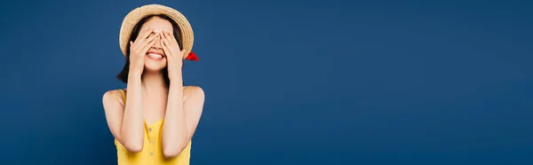 Smiling girl in straw hat with hands on eyes isolated on blue — Stock Photo