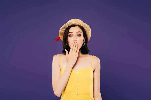 Шокированная девушка в соломенной шляпе, покрывающей рот рукой, изолированной на фиолетовый — стоковое фото