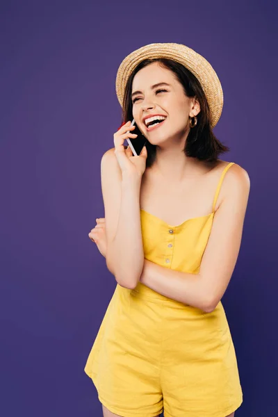 Chica feliz en sombrero de paja hablando en el teléfono inteligente y riendo aislado en púrpura - foto de stock