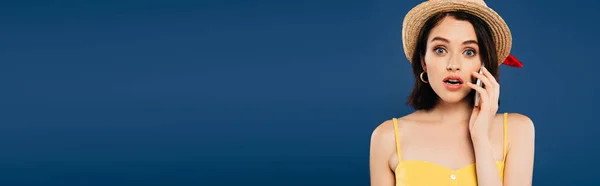 Panoramic shot of shocked girl in straw hat talking on smartphone isolated on blue — Stock Photo