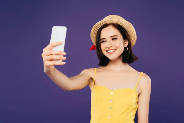 Улыбающаяся девушка в соломенной шляпе делает селфи на смартфоне, изолированном на фиолетовый — стоковое фото
