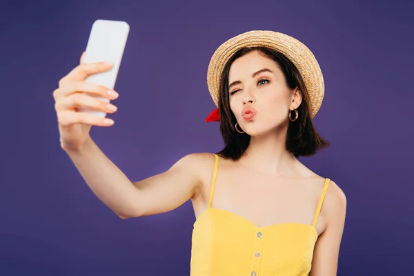 Smiling girl in straw hat pouting lips while taking selfie on smartphone isolated on purple — Stock Photo