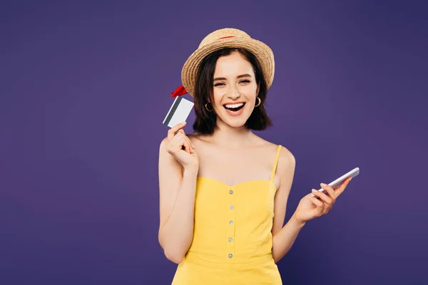 Chica sonriente en sombrero de paja celebración de teléfono inteligente y tarjeta de crédito aislado en púrpura - foto de stock