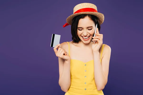 Усміхнена дівчина в солом'яному капелюсі говорить на смартфоні і тримає кредитну картку ізольовано на фіолетовому — стокове фото