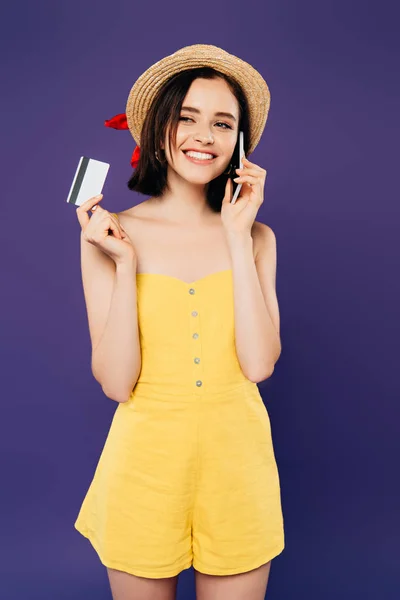 Улыбающаяся девушка в соломенной шляпе разговаривает по смартфону и держит кредитку изолированной на фиолетовый — стоковое фото