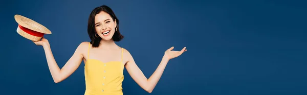 Plano panorámico de niña sonriente sosteniendo sombrero de paja aislado en azul - foto de stock