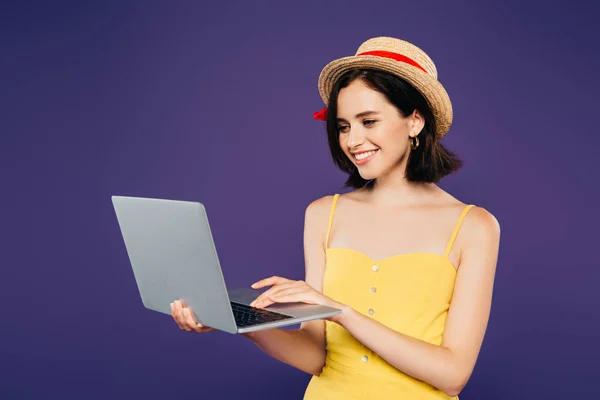 Chica sonriente en sombrero de paja usando portátil aislado en púrpura - foto de stock