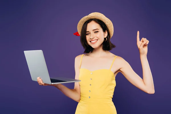 Smiling girl in straw hat holding laptop and showing idea gesture isolated on purple — Stock Photo