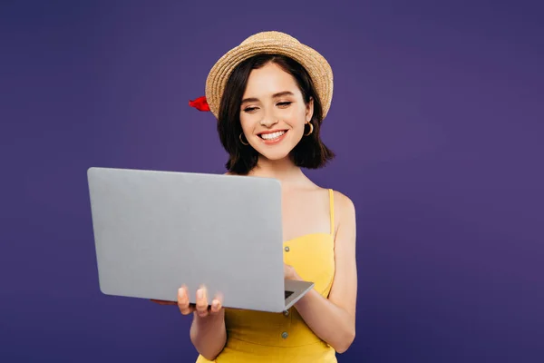 Chica sonriente en sombrero de paja usando portátil aislado en púrpura - foto de stock