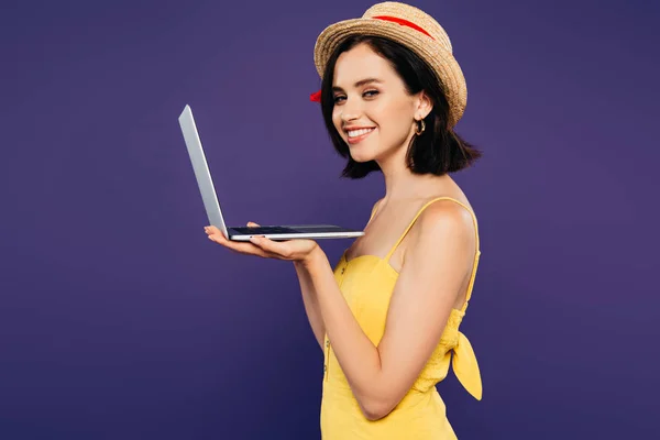 Vista lateral de chica sonriente en sombrero de paja sosteniendo portátil aislado en púrpura - foto de stock