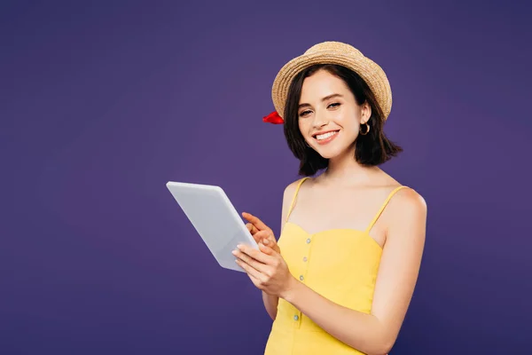 Улыбающаяся красивая девушка в соломенной шляпе, держащая цифровой планшет изолирован на фиолетовый — стоковое фото