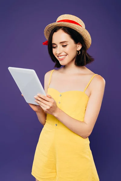 Sonriente chica bonita en sombrero de paja sosteniendo tableta digital aislado en púrpura - foto de stock