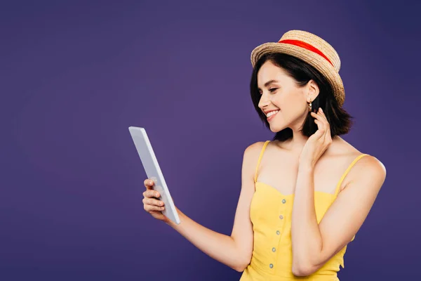 Sonriente chica bonita en sombrero de paja sosteniendo tableta digital aislado en púrpura - foto de stock