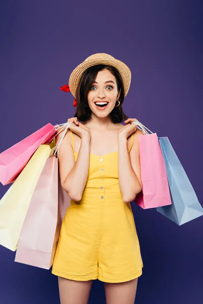 Chica sonriente excitada en sombrero de paja con bolsas de compras aisladas en púrpura - foto de stock