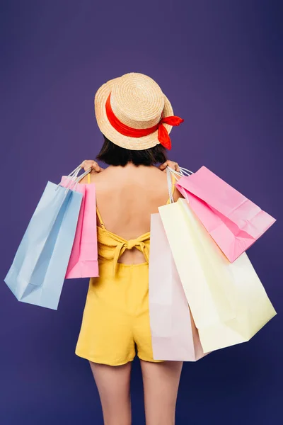 Vista trasera de chica en sombrero de paja con bolsas de compras aisladas en púrpura - foto de stock