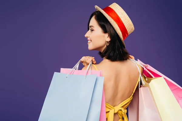 Vista posteriore di ragazza sorridente in cappello di paglia con borse della spesa isolate su viola — Foto stock