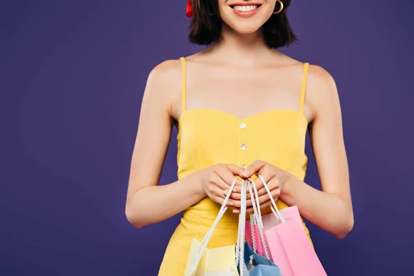 Частковий вид усміхненої дівчини в солом'яному капелюсі з сумками для покупок ізольовано на фіолетовому — стокове фото