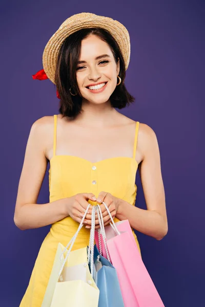 Ragazza sorridente in cappello di paglia che tiene borse della spesa isolate su viola — Foto stock