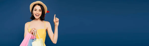 Panoramic shot of excited smiling girl in straw hat with shopping bags showing idea sign isolated on blue — Stock Photo