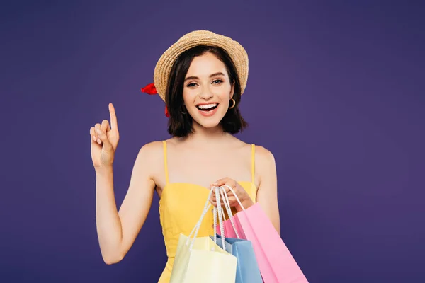 Chica sonriente excitada en sombrero de paja con bolsas de compras que muestran idea signo aislado en púrpura - foto de stock