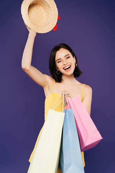 Smiling girl with shopping bags holding straw hat isolated on purple — Stock Photo