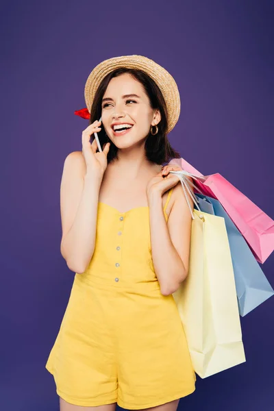 Ragazza sorridente in cappello di paglia con borse della spesa parlare su smartphobne isolato su viola — Foto stock