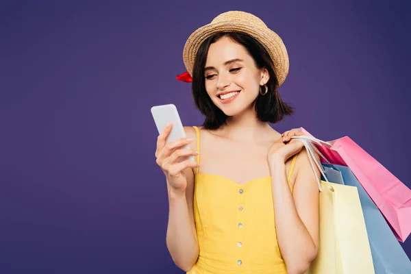 Smiling girl in straw hat with shopping bags using smartphone isolated on purple — Stock Photo