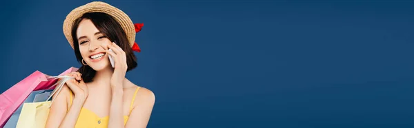 Foto panoramica di ragazza sorridente in cappello di paglia con shopping bag che parla su smartphone isolato su blu — Foto stock