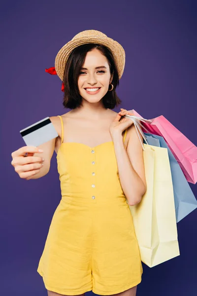 Chica sonriente en sombrero de paja con bolsas de compras presentando tarjeta de crédito aislada en púrpura - foto de stock