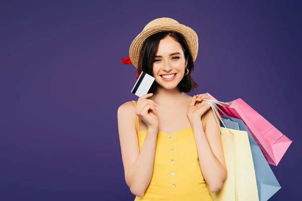 Ragazza sorridente in cappello di paglia con borse della spesa in possesso di carta di credito isolata su viola — Foto stock