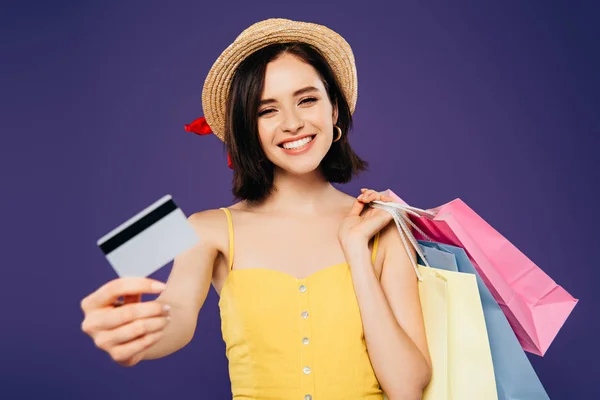 Fille souriante dans un chapeau de paille avec des sacs à provisions montrant carte de crédit isolé sur violet — Photo de stock