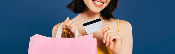 Plano panorámico de chica sonriente con bolsas de compras y tarjeta de crédito aislada en azul - foto de stock