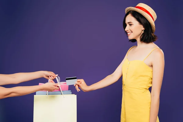 Усміхнена дівчина в солом'яному капелюсі бере сумки і дає кредитну картку ізольовано на фіолетовому — стокове фото