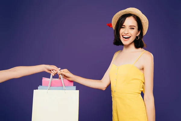 Sorridente ragazza in cappello di paglia prendere shopping bags isolato su viola — Stock Photo