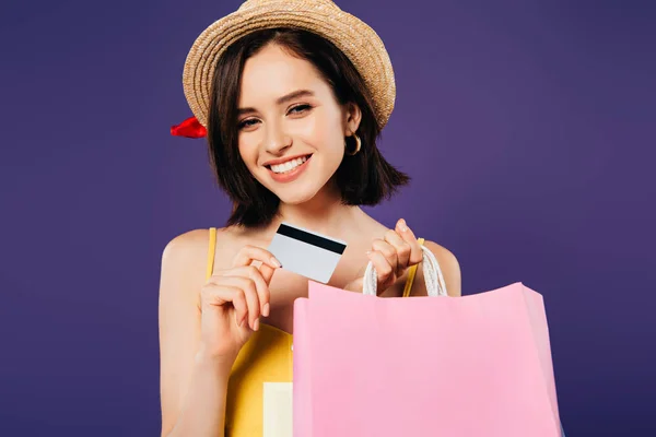 Chica sonriente en sombrero de paja con bolsas de compras y tarjeta de crédito aislada en púrpura - foto de stock