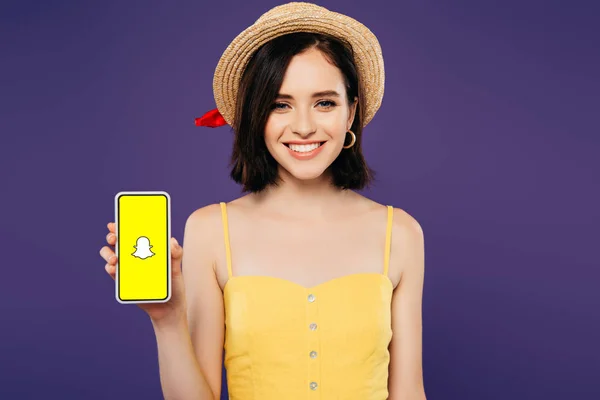 KYIV, UKRAINE - 3 JUILLET 2019 : fille souriante en chapeau de paille tenant smartphone avec application Snapchat isolé sur violet — Photo de stock