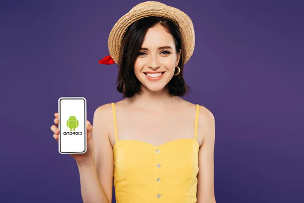 KYIV, UKRAINE - 3 JUILLET 2019 : fille souriante en chapeau de paille tenant smartphone avec logo Android isolé sur violet — Photo de stock