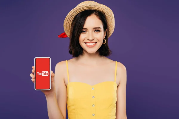 КИЕВ, Украина - 3 июля 2019 года: улыбающаяся девушка в соломенной шляпе держит смартфон с приложением youtube изолированным на фиолетовый — стоковое фото