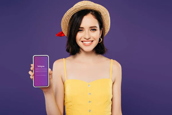 KYIV, UCRANIA - 3 de julio de 2019: niña sonriente con sombrero de paja sosteniendo el teléfono inteligente con aplicación Instagram aislada en púrpura - foto de stock