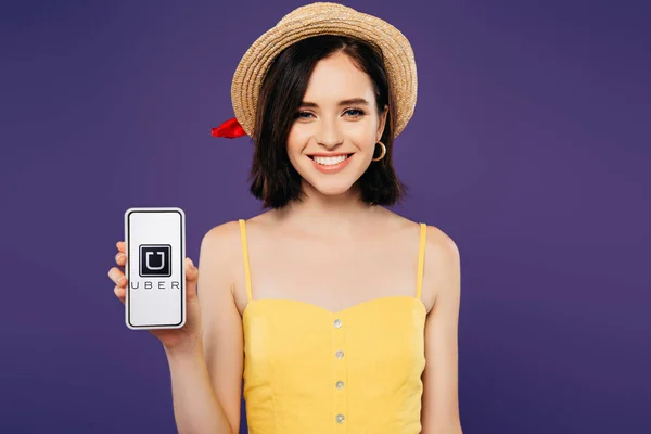 KYIV, UCRAINA - 3 LUGLIO 2019: ragazza sorridente in cappello di paglia con smartphone in mano con app uber isolata su viola — Foto stock