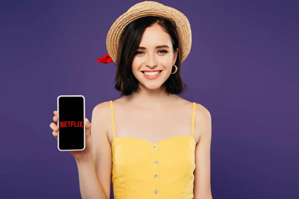 Київ, Україна-3 липня 2019: усміхнена дівчина в солом'яному капелюсі тримає смартфон з додатком Netflix, ізольованим на фіолетовому — стокове фото