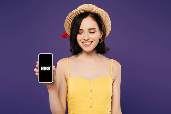 KYIV, UKRAINE - JULHO 3, 2019: menina sorridente em chapéu de palha segurando smartphone com aplicativo HBO isolado em roxo — Fotografia de Stock