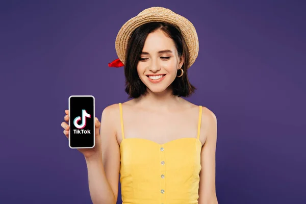 KYIV, UKRAINE - 3 JUILLET 2019 : fille souriante en chapeau de paille tenant smartphone avec application tiktok isolée sur violet — Photo de stock