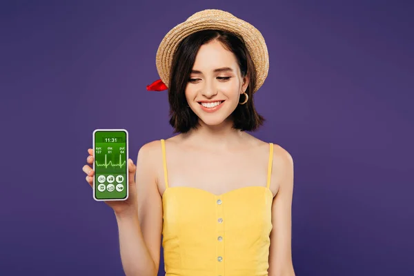 Улыбающаяся девушка в соломенной шляпе, держащая смартфон с приложением для здравоохранения, изолированным на фиолетовый — стоковое фото