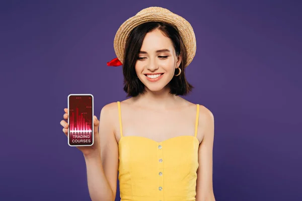 Усміхнена дівчина в солом'яному капелюсі тримає смартфон із додатком торгових курсів ізольовано на фіолетовому — стокове фото