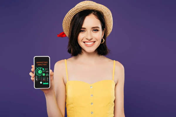 Chica sonriente en sombrero de paja celebración de teléfono inteligente con aplicación de análisis de marketing aislado en púrpura - foto de stock