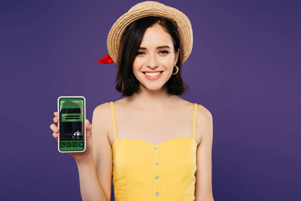 Chica sonriente en sombrero de paja celebración de teléfono inteligente con aplicación de reserva aislado en púrpura - foto de stock
