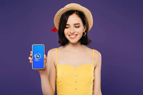 Київ, Україна-3 липня 2019: усміхнена дівчина в солом'яному капелюсі тримає смартфон з додатком Shazam, ізольованим на фіолетовому — стокове фото
