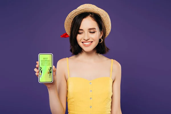 Chica sonriente en sombrero de paja celebración de teléfono inteligente con la aplicación de compras beat aislado en púrpura - foto de stock